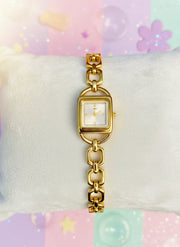 Gold link bracelet watch with square face on white cushion, colorful pastel background
