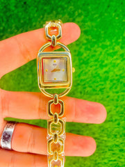 Gold women's midi watch with square dial and linked bracelet held over green background