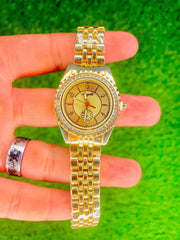 Luxury gold women's wristwatch with stone-studded bezel and linked bracelet on green background