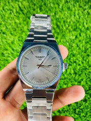Tissot PRX men's stainless steel watch with silver dial and date window held on hand over green grass
